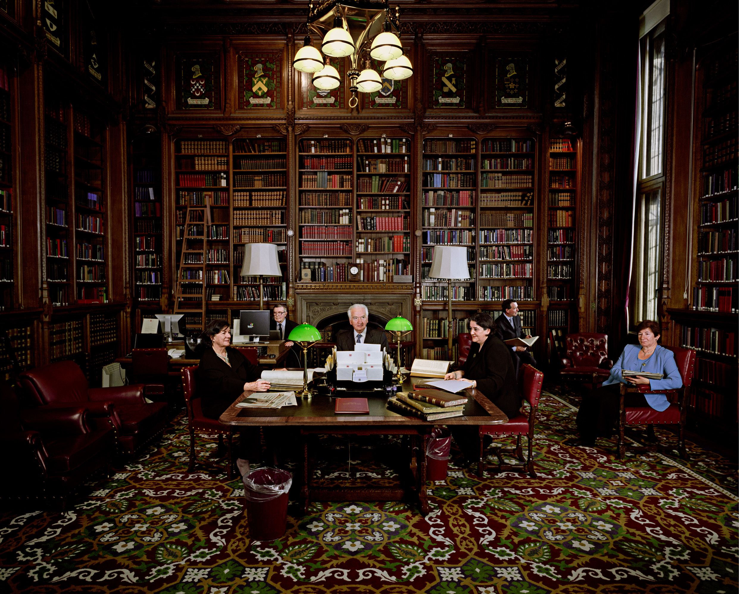 A portrait photo of members in the House of Lords Library. There are book shelves surround them, with a large wooden table at the centre of the image where Lord Mackay is seated.