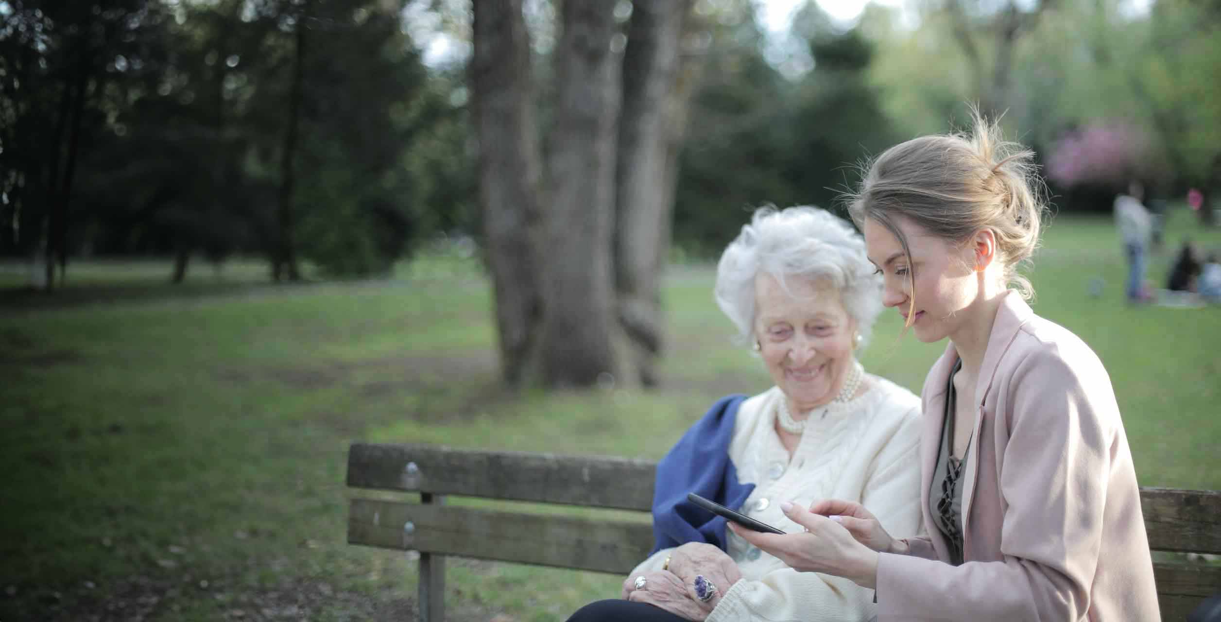 Image of an elderly lady and a young woman sitting on a bench, looking at a mobile phone.