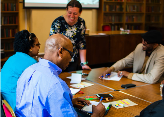 A facilitator stands in front of a small group of adults sat around a table, laughing with them
