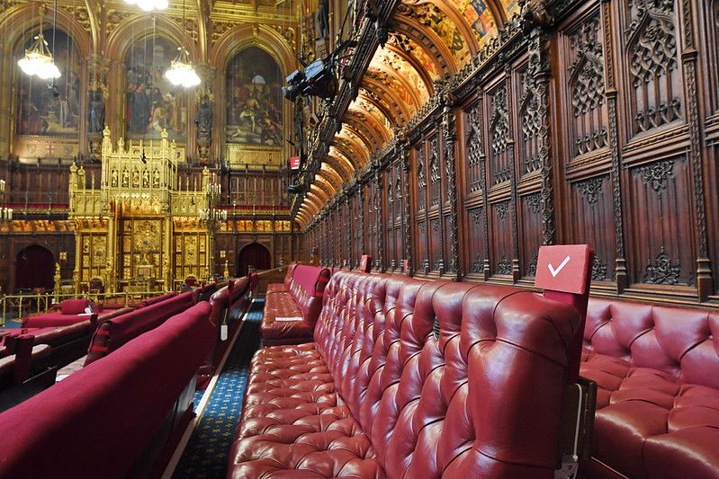 An image from the seats on the left hand side (opposition side) of the Lords Chamber, facing the throne which is visible in the background, below a large gold canopy. The seats in the foreground are red leather benches. There is ornate dark panelling behind them, with decorative shields painted above.