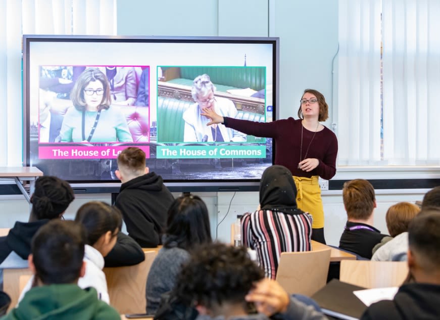A facilitator wearing a yellow skirt stands in front of a room full of people, gesturing to a screen on which images of someone speaking in the House of Commons and someone speaking in the House of Lords