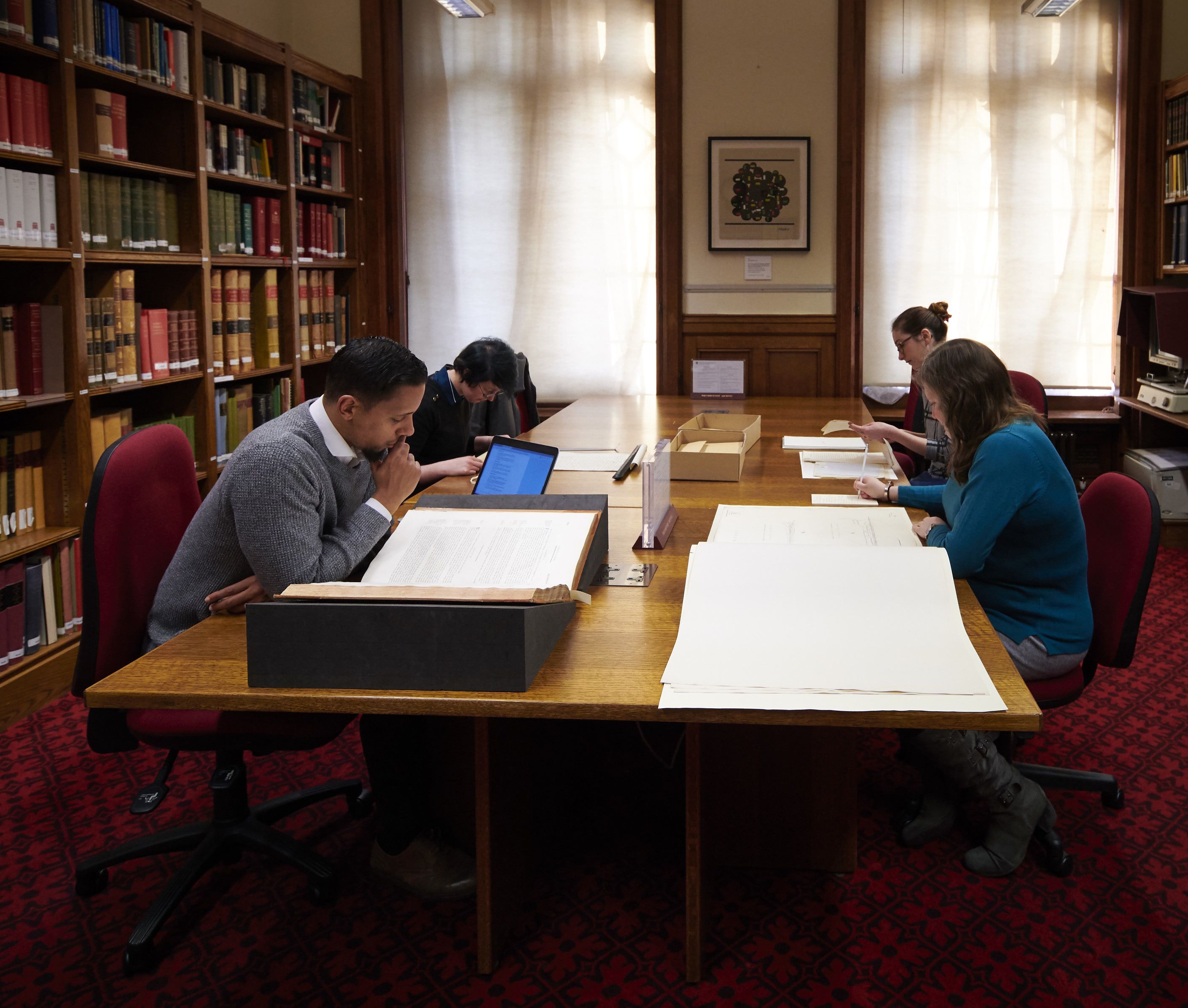Colour photograph of an archive search room. There are four researchers looking at paper documents on a large wooden desk.