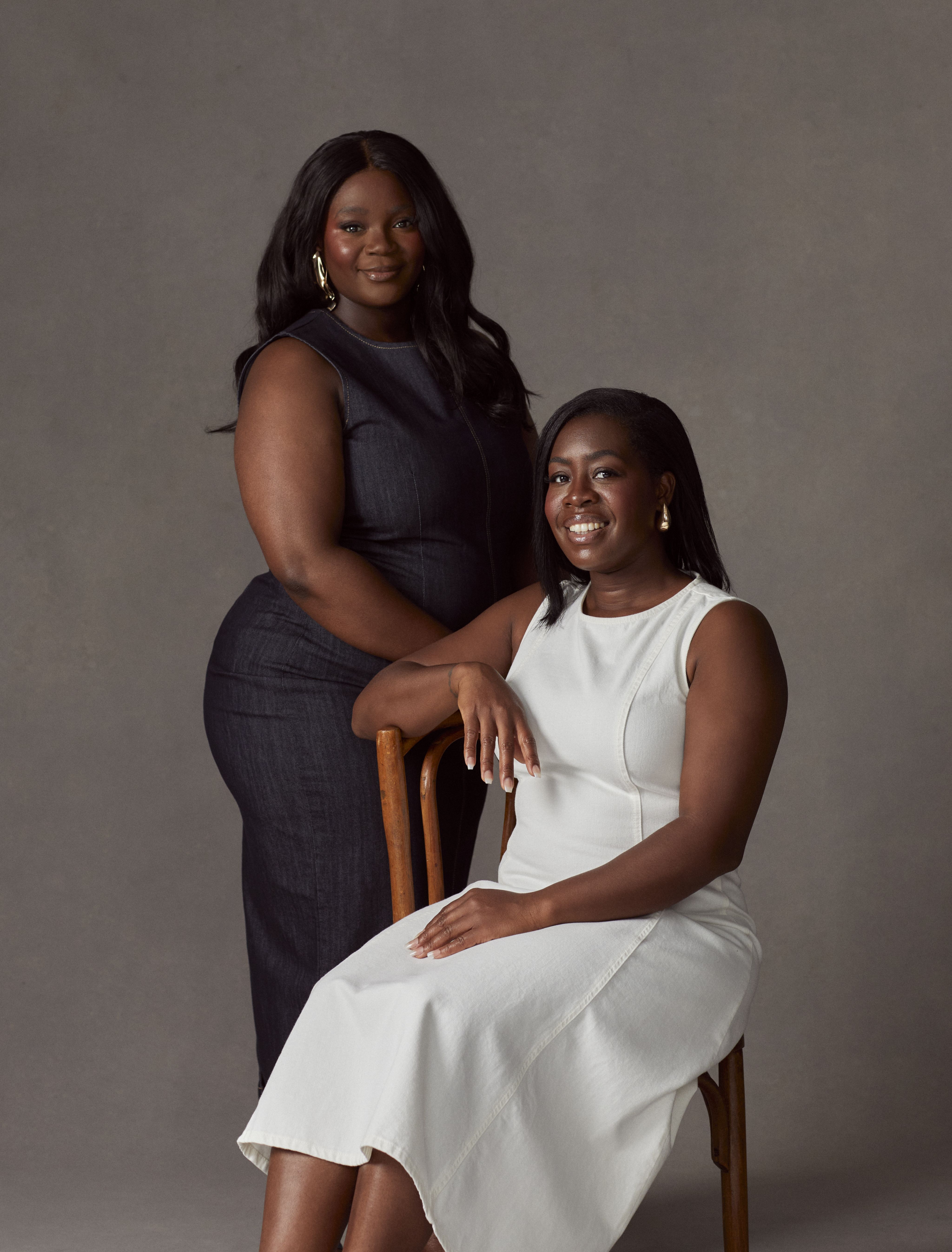 Two black women face the camera smiling, one is sat on a chair wearing a white dress and the other is stood beside her wearing a long navy dress.