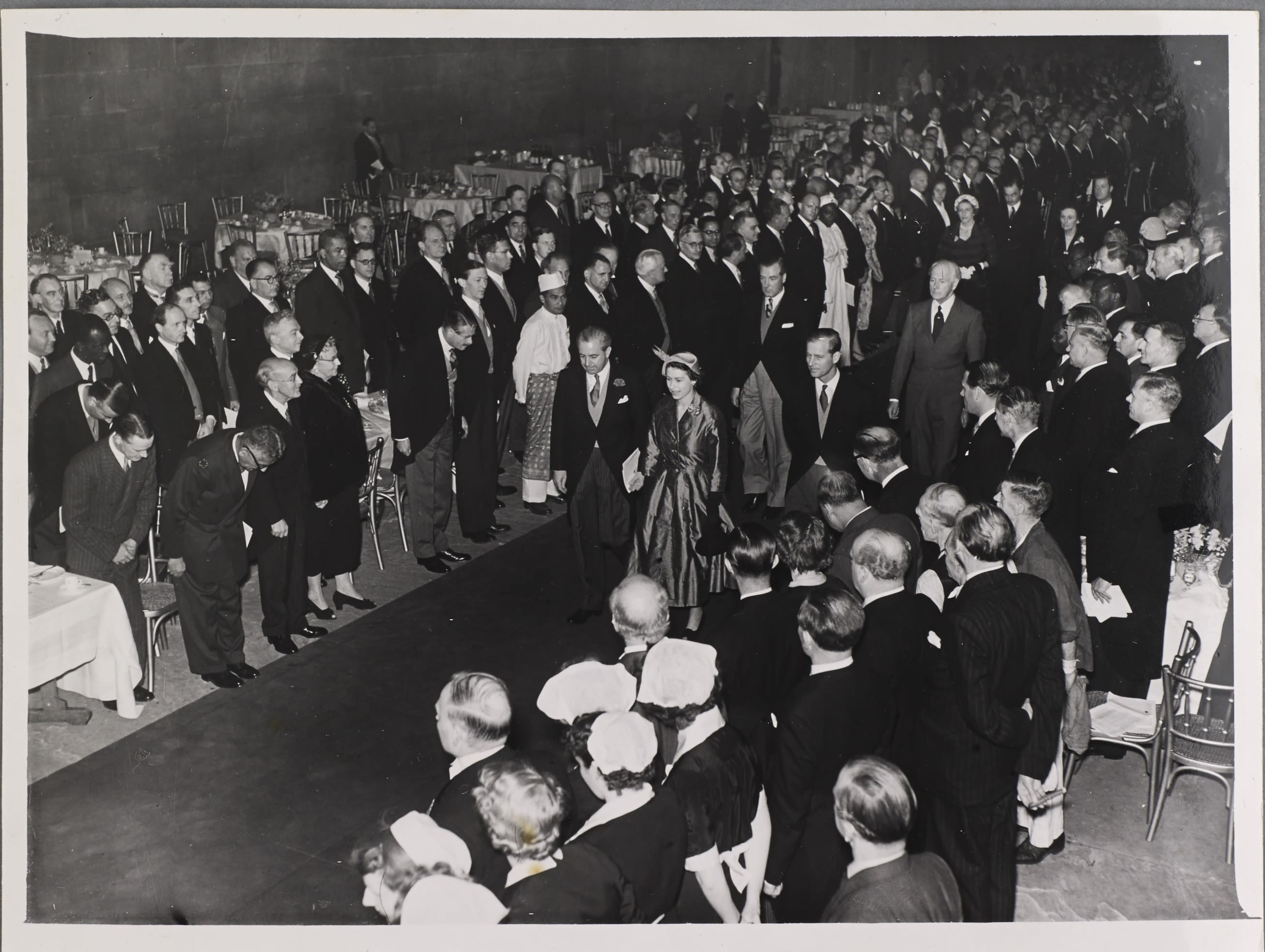 Black and white photograph showing Queen Elizabeth II walking down a carpet with people either side looking on.
